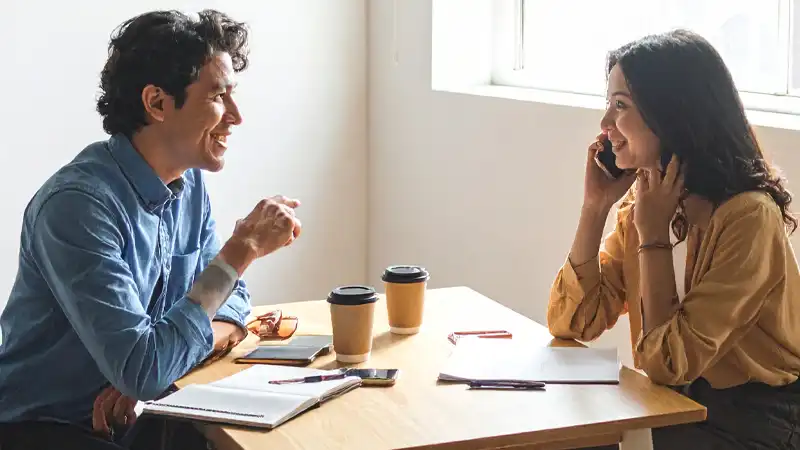 Two colleagues chat across a wooden table in a bright room, with notebooks, phones, and coffee cups on the table.